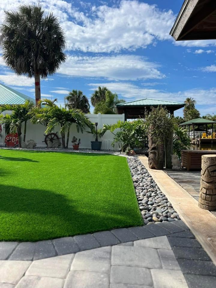 Artificial turf lawn with river rock border and paver walkway on a waterfront lot with tiki hut